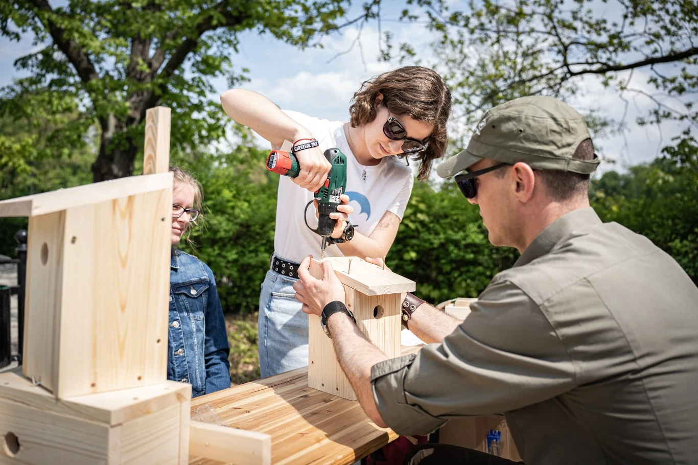 Dwójka dorosłych i dziecko uczestniczą w warsztatach budowania drewnianych budek dla ptaków. Kobieta używa wiertarki, by połączyć elementy, podczas gdy mężczyzna trzyma konstrukcję. Dziecko przygląda się z zainteresowaniem.