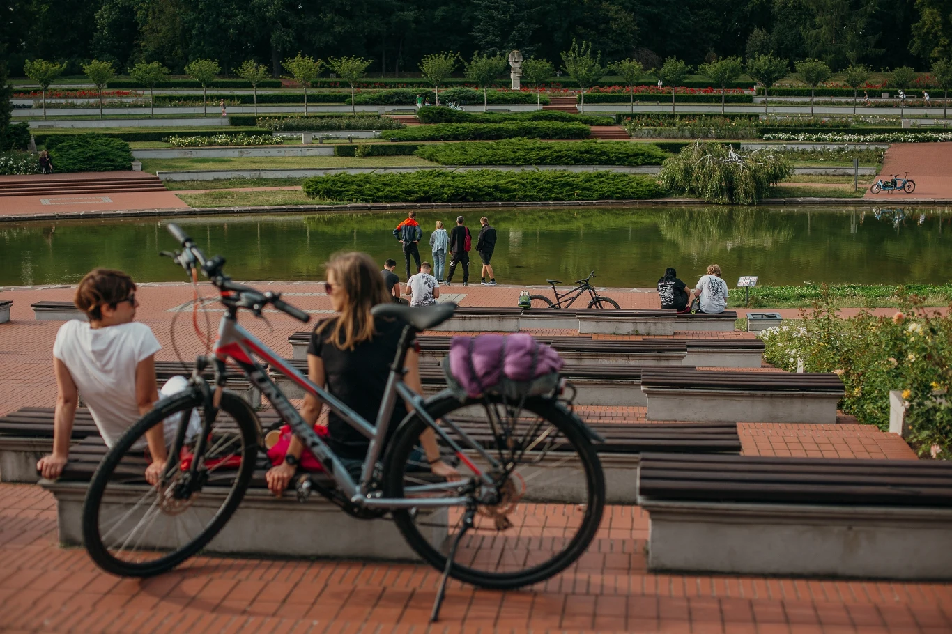 Ludzie odpoczywający w parku nad niewielkim stawem. Na pierwszym planie widoczna jest dziewczyna siedząca obok roweru. W tle znajduje się zielona roślinność oraz kilka osób spacerujących w oddali.