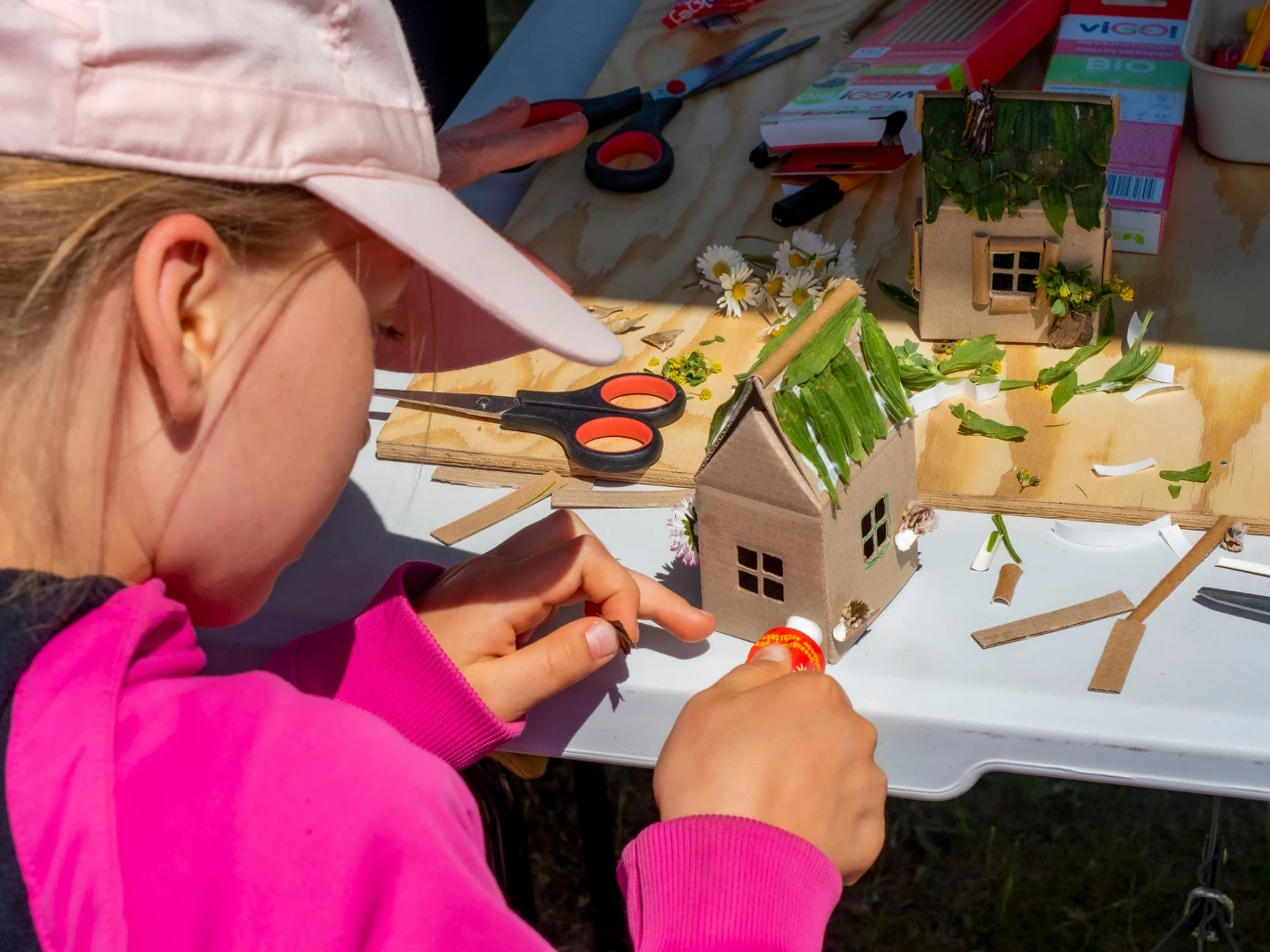 Dziecko w różowej kurtce i czapce tworzy makietę małego domku przy stoliku. Domek ozdobiony jest zielonymi i białymi roślinami. Obok leżą nożyczki oraz inne narzędzia do prac plastycznych.