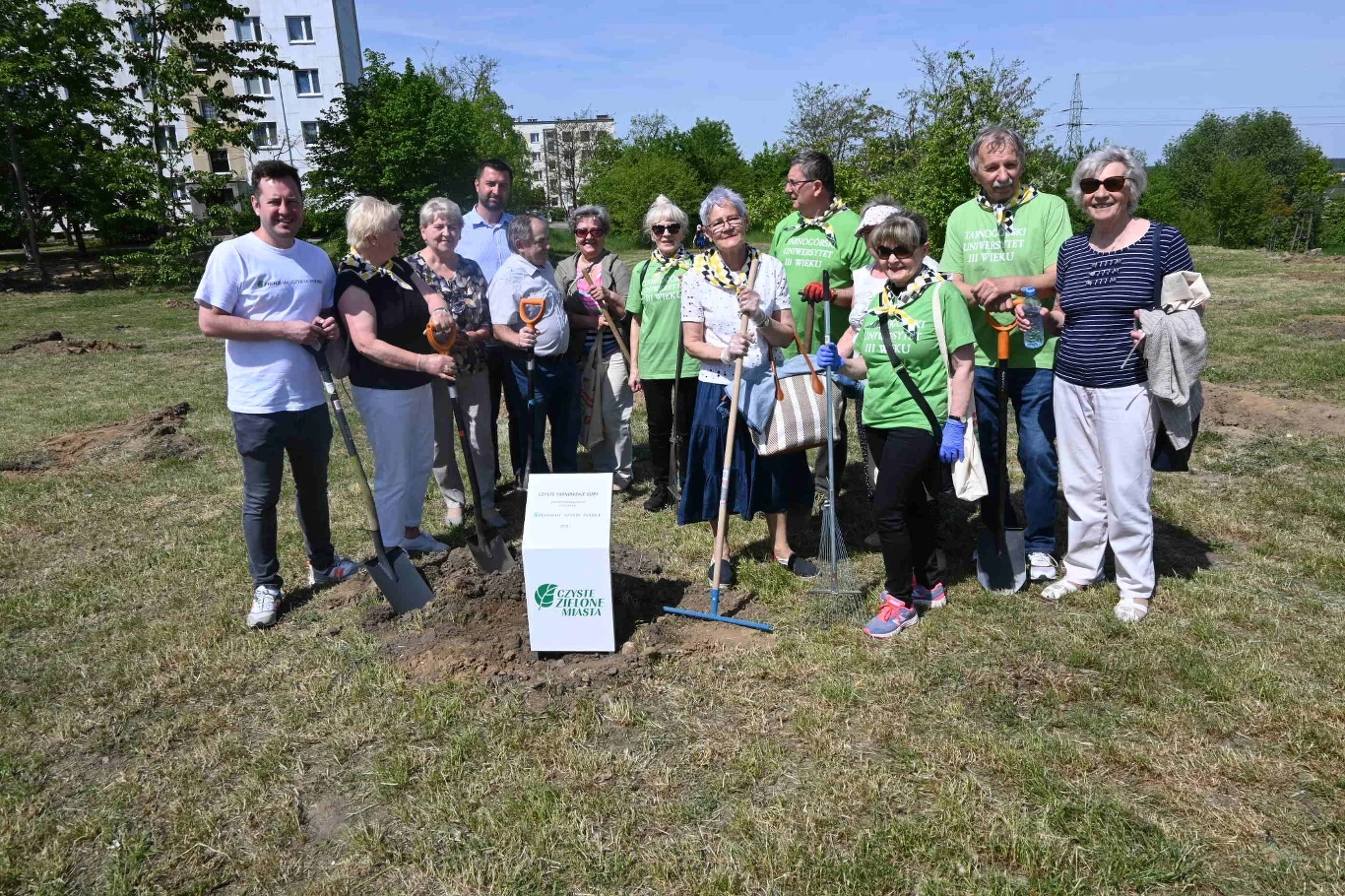 Czyste Zielone Miasta fot. UM Tarnowskie Góry Grupa osób w różnym wieku pozuje na trawniku podczas wydarzenia związanego z ekologią. Niektórzy trzymają narzędzia ogrodnicze, na środku widoczna jest tabliczka z logo projektu. W tle widoczne są drzewa i budynki mieszkalne.