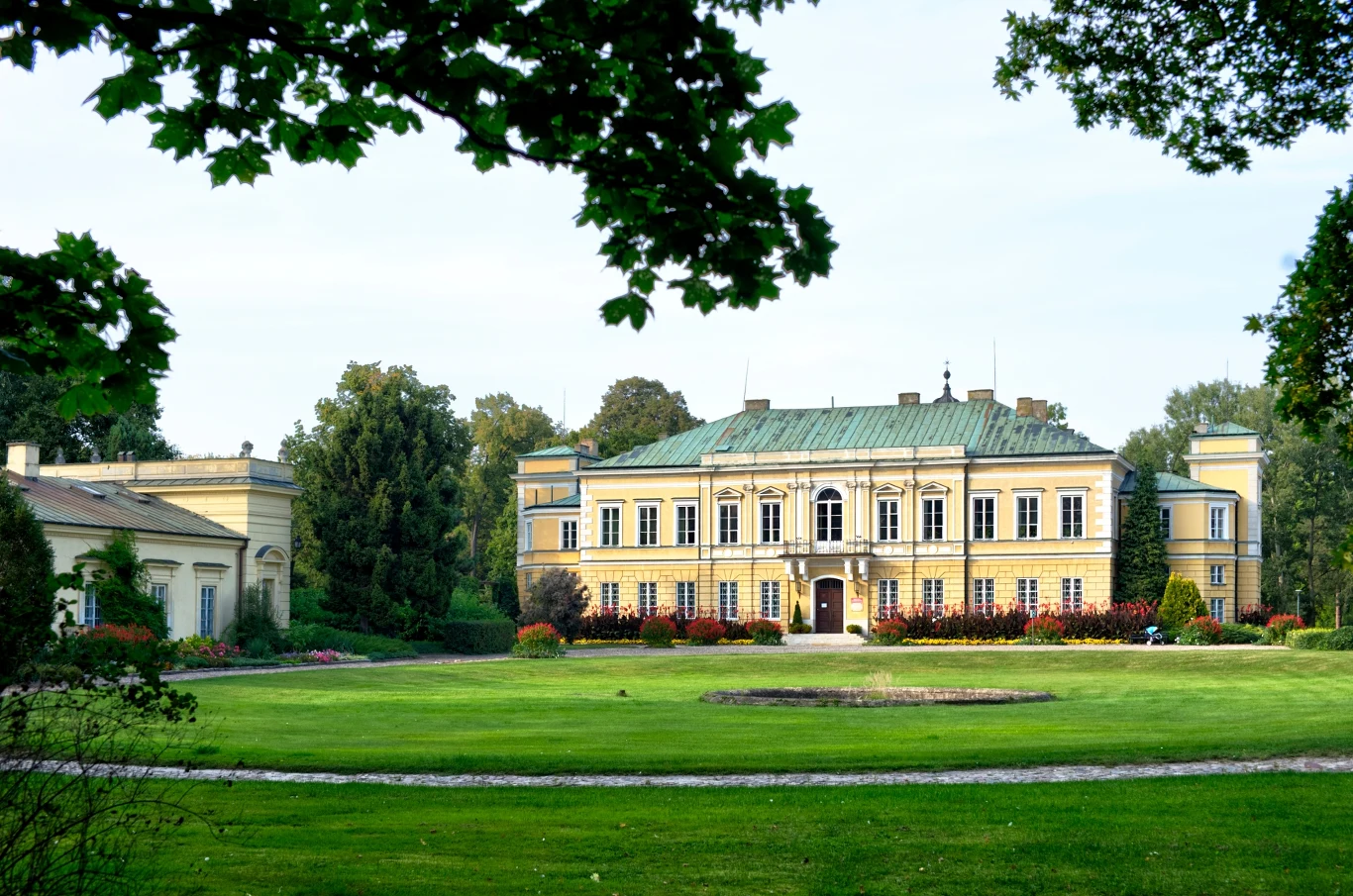 Czyste Zielone Miasta fot. UM Skierniewice Zabytkowy pałac otoczony pięknym ogrodem i zielenią. Budynek jest dwukondygnacyjny, z charakterystycznym zielonym dachem. W tle widoczne są drzewa i krzewy, a na pierwszym planie trawnik.