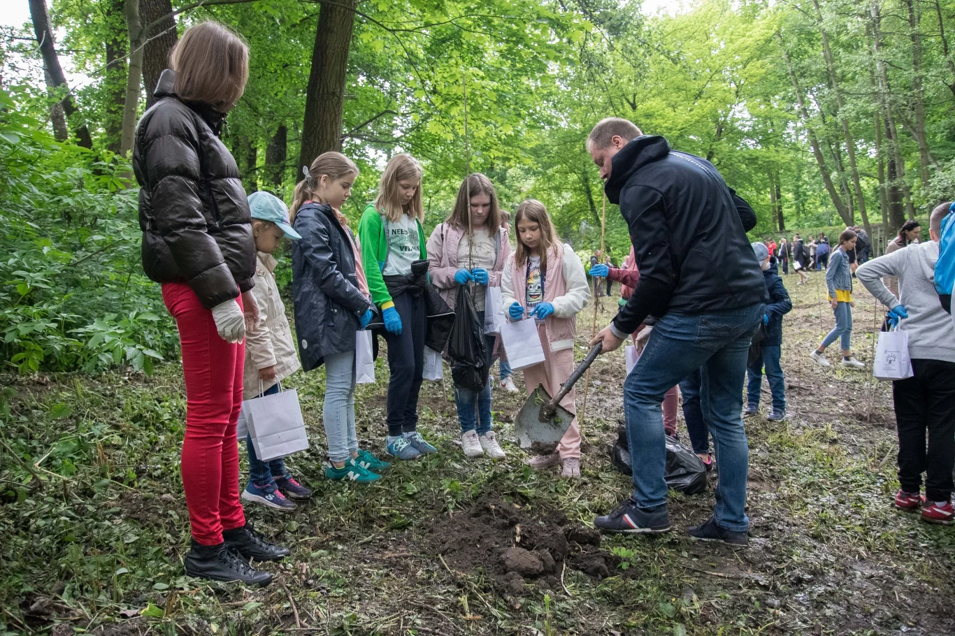 Czyste Zielone Miasta fot. UM Skierniewice Grupa dzieci i dorosły uczestniczą w akcji sprzątania w lesie. Dorośli instruują dzieci, jak podnosić śmieci z ziemi. Wszyscy mają na sobie rękawiczki ochronne, a wokół panuje atmosfera współpracy i edukacji ekologicznej.
