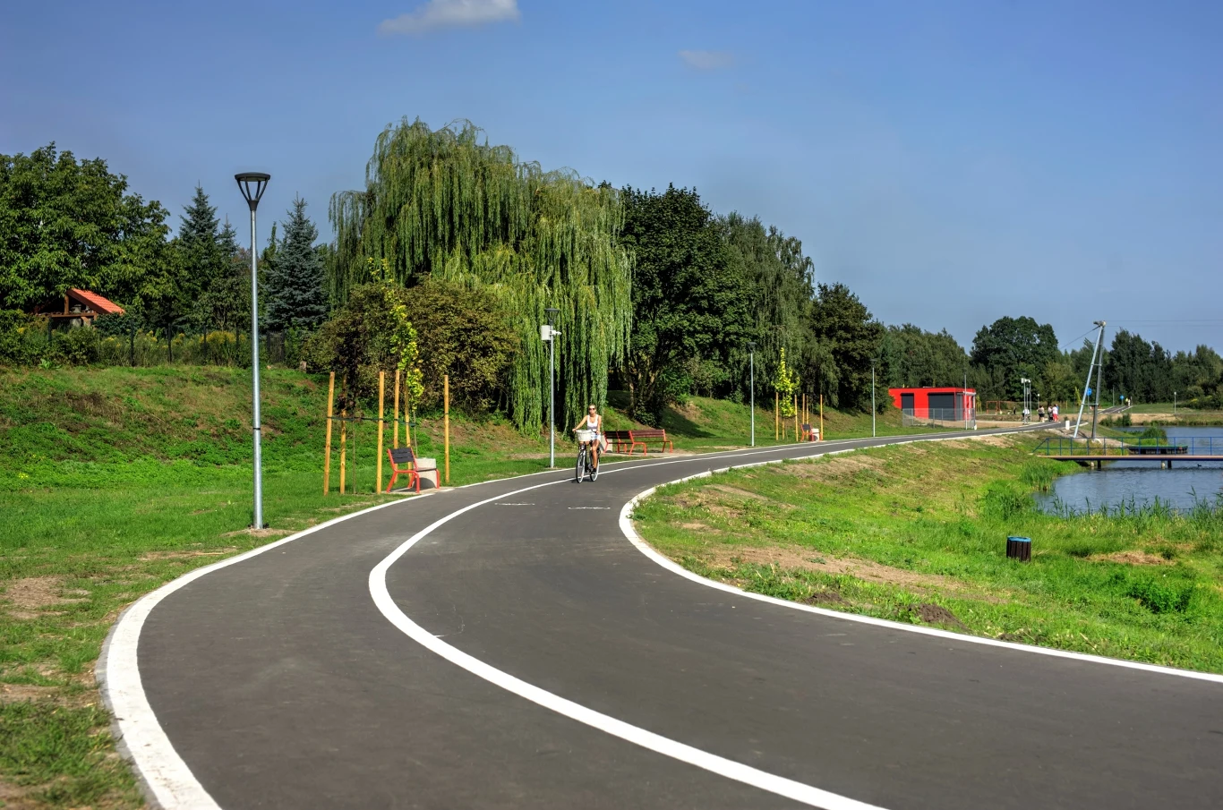Czyste Zielone Miasta fot. UM Skierniewice Asfaltowa ścieżka rowerowa biegnąca przez zielony park, z drzewami i krzewami po bokach. Rowerzysta na rowerze jedzie w oddali, a przy ścieżce znajdują się ławki. Po prawej stronie widoczny jest staw z czerwoną altaną.