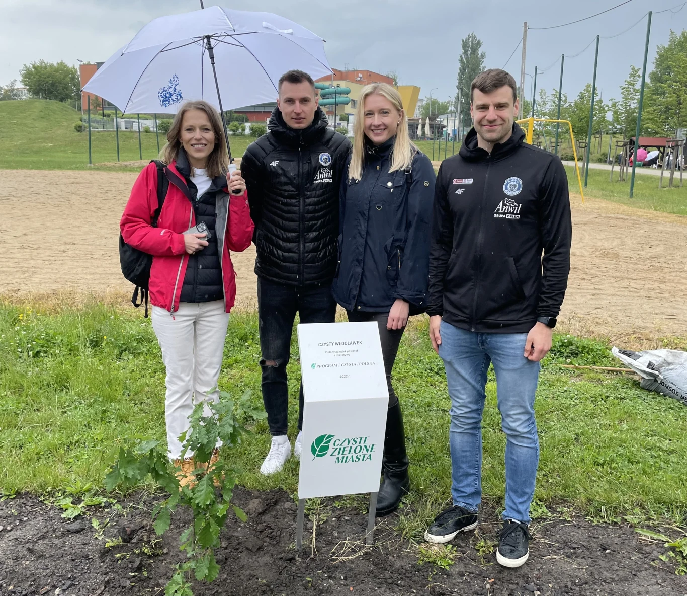 Grupa czterech osób stoi na trawie przy tabliczce z napisem, jeden z nich trzyma parasol. W tle widać boisko i drzewa.