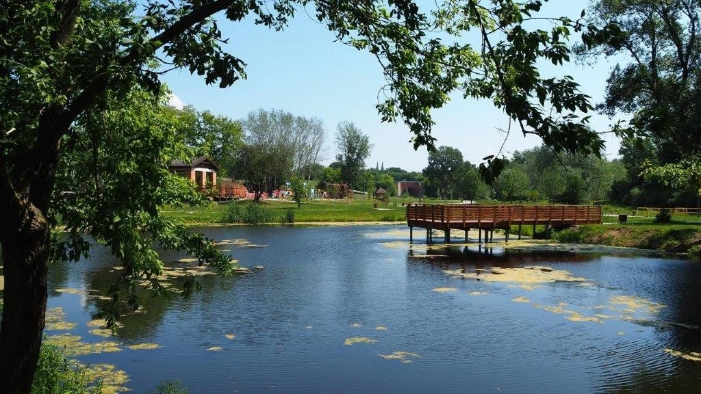Spokojny krajobraz z jeziorem otoczonym drzewami, pomostem na wodzie oraz domkami i zielenią w tle. Odbicia drzew w wodzie dodają urokliwego klimatu.