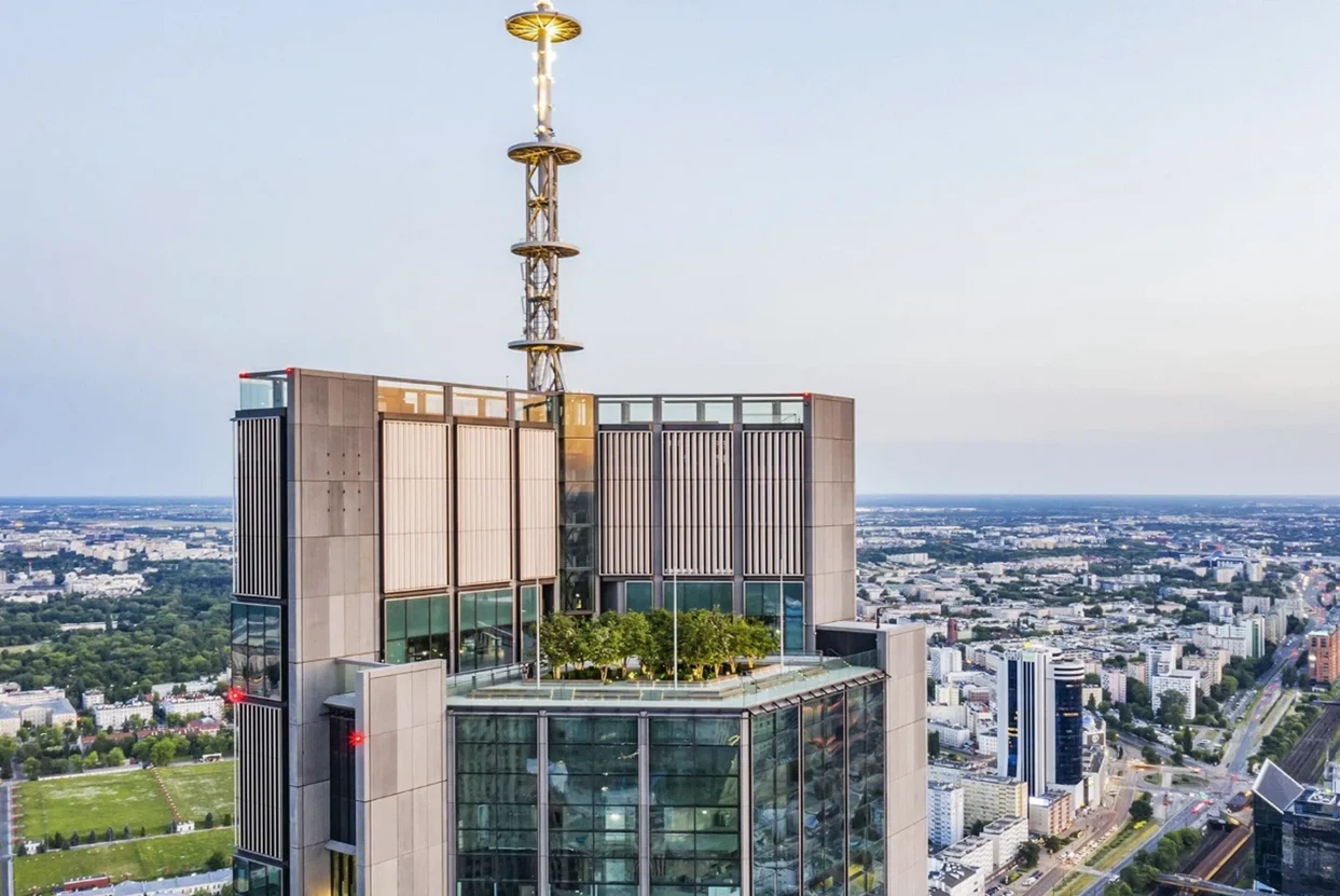 Tarasy i bar znajdą się ponad 200 metrów nad ziemią Widok na nowoczesny wieżowiec z anteną na dachu, otoczony miastem. Na szczycie budynku znajduje się taras z zielenią. W tle rozciąga się panorama miasta z ulicami i budynkami.