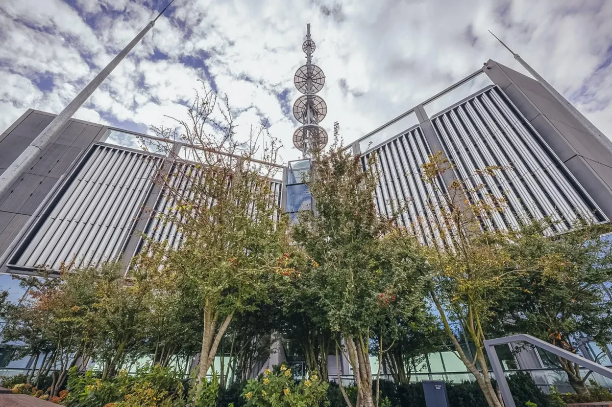 Dolny taras będzie obsadzony drzewami Nowoczesny budynek z dużą anteną satelitarną na dachu, otoczony drzewami, pod niebem pełnym chmur. Budowla ma geometryczne kształty i przeszklone elementy.