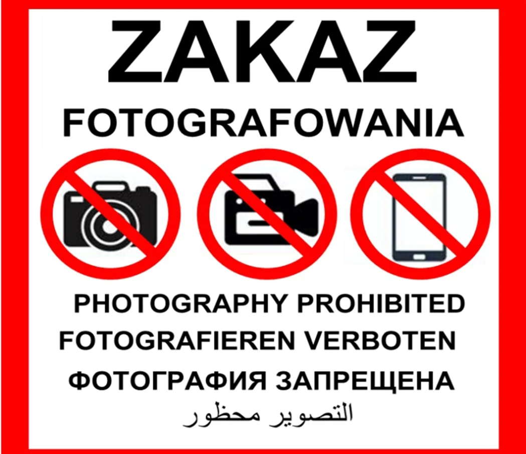 Znak zakazu fotografowania przedstawiający ikony aparatu, kamery i smartfona z przekreśleniami. Zawiera napisy w sześciu językach, informujące o zakazie robienia zdjęć.