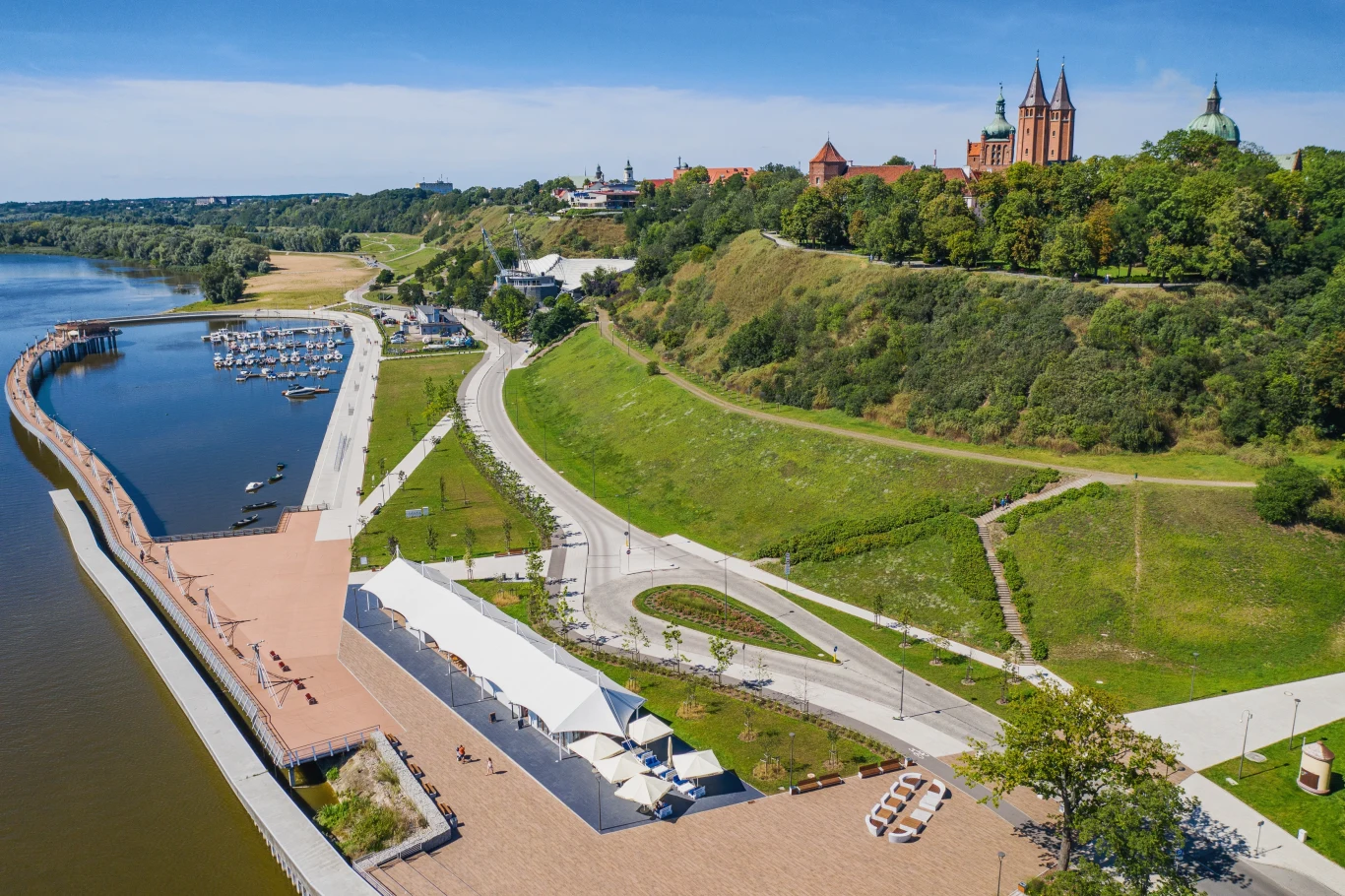 Czyste Zielone Miasta fot. UM Płock Panorama nadbrzeża z widokiem na zamek na wzgórzu, promenadę i marinę przy rzece w pogodny dzień.