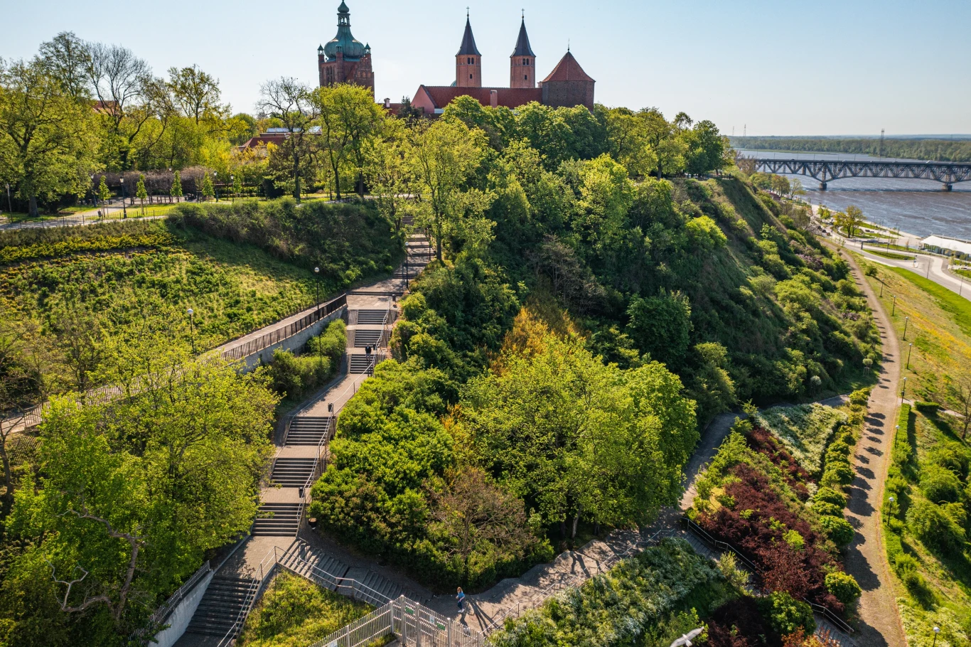 Czyste Zielone Miasta fot. UM Płock Widok na wzgórze z zamkiem otoczony bujną zielenią, schody prowadzące na szczyt oraz rozległa panorama na rzekę i most. Na pierwszym planie roślinność o różnorodnej strukturze, a w tle zabytkowe budowle.