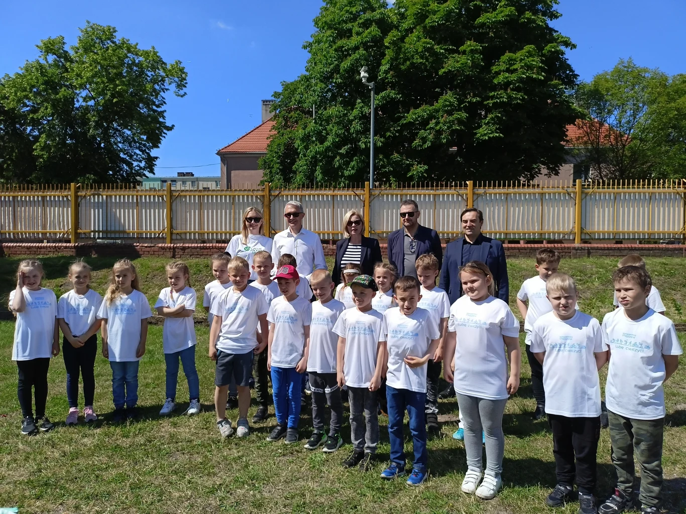 Grupa dzieci w białych koszulkach oraz kilku dorosłych stoi na trawie, prawdopodobnie podczas wydarzenia szkolnego. W tle znajduje się ogrodzenie oraz drzewa.
