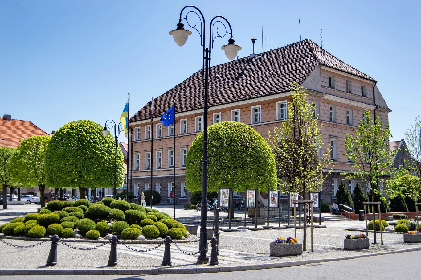 Budynek administracyjny otoczony zadbaną zielenią i flagami na placu miejskim, z widocznym niebieskim niebem.