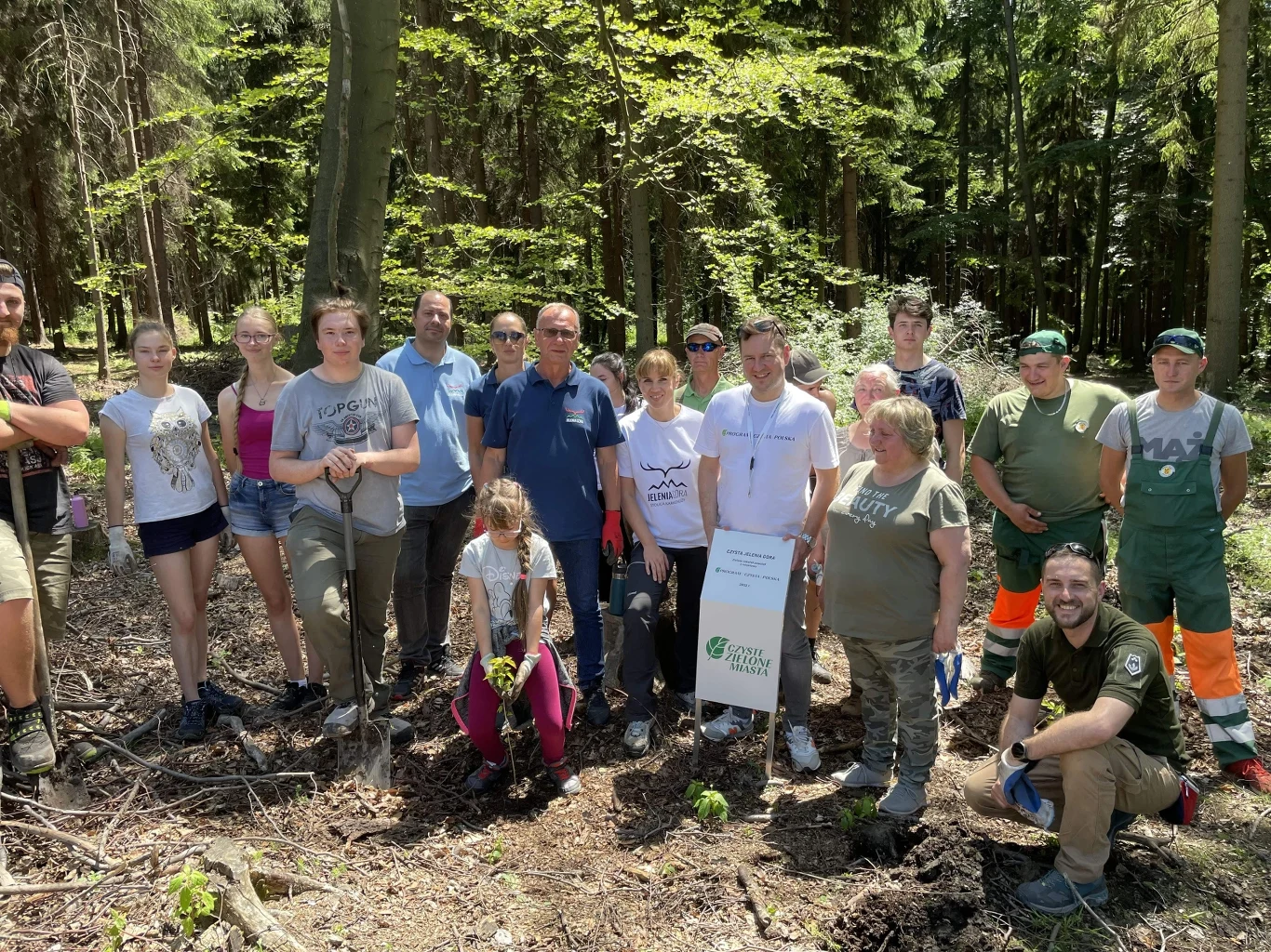 Grupa ludzi pośród drzew w lesie, część z nich trzyma narzędzia ogrodnicze. W centralnej części tabliczka z napisem sugerującym udział w akcji związanej z ekologią.