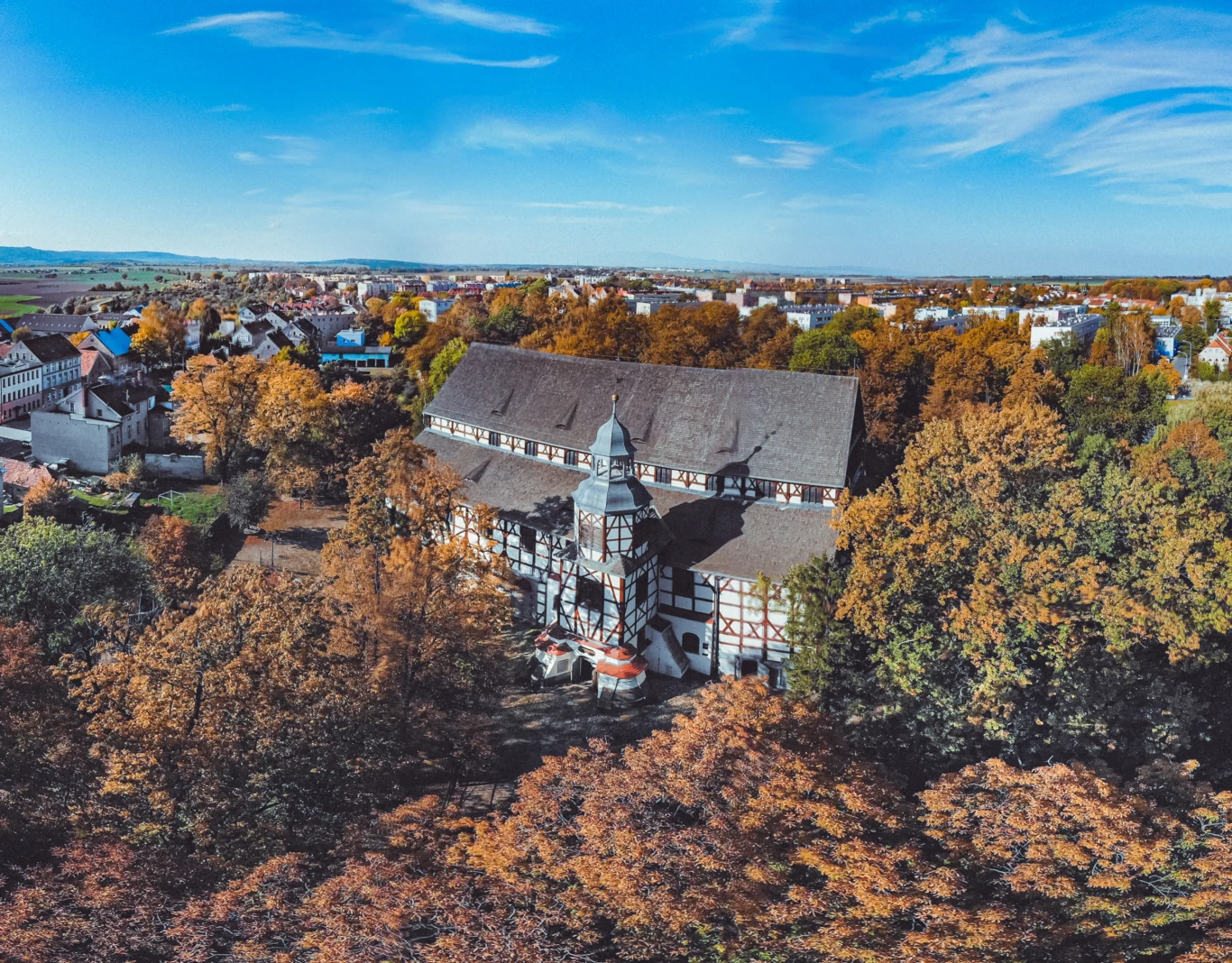 Panoramiczny widok na Kościół Pokoju w Świdnicy otoczony jesiennymi drzewami. Charakterystyczna szachulcowa architektura budynku wyróżnia się na tle różnorodnego krajobrazu miejskiego.