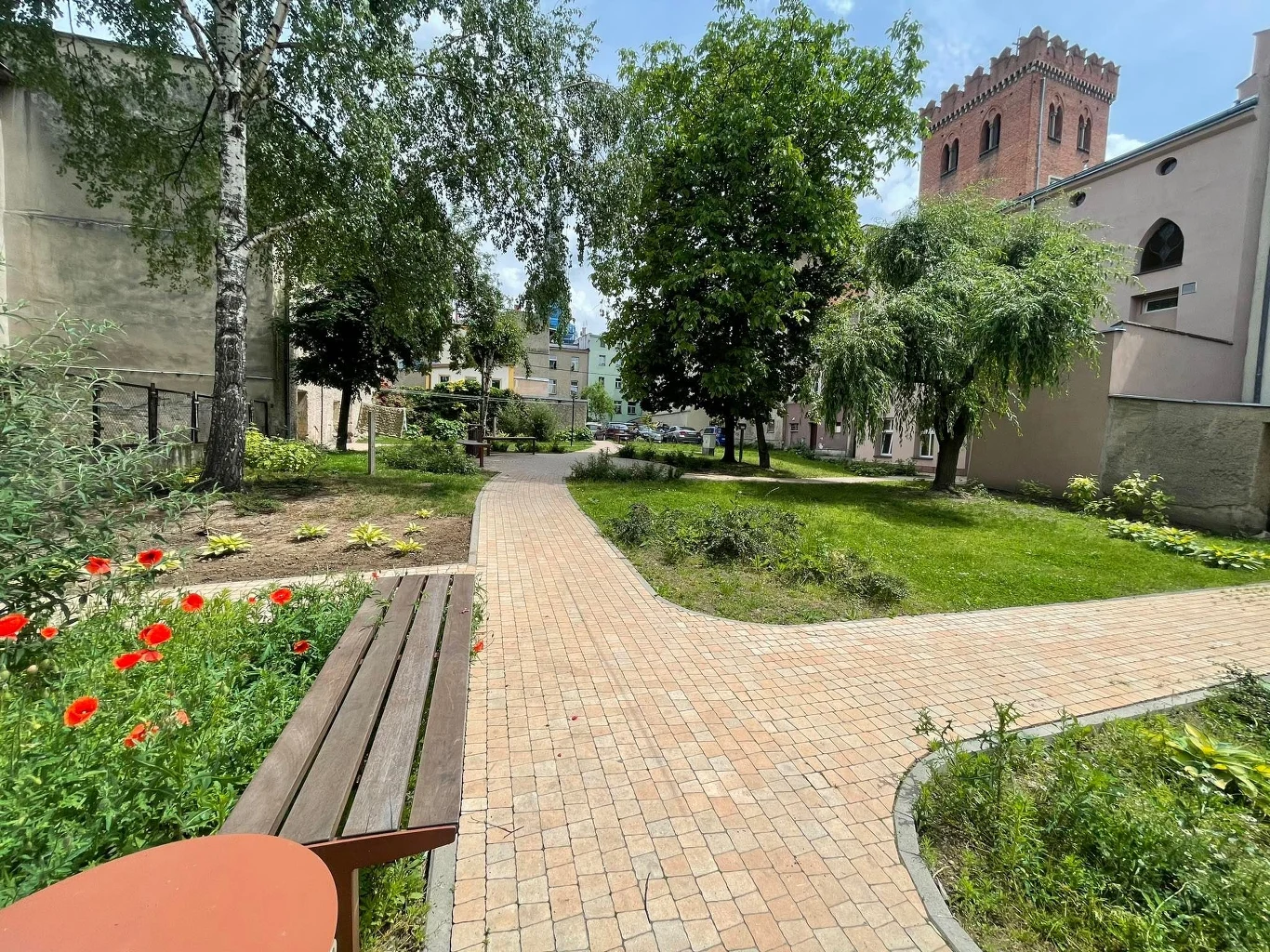 Ogród miejski z brukowaną ścieżką prowadzącą między drzewami i kwiatami. W tle widać zabytkową ceglaną wieżę i kilka budynków mieszkalnych. Na pierwszym planie znajduje się drewniana ławka oraz kwitnące maki po lewej stronie.