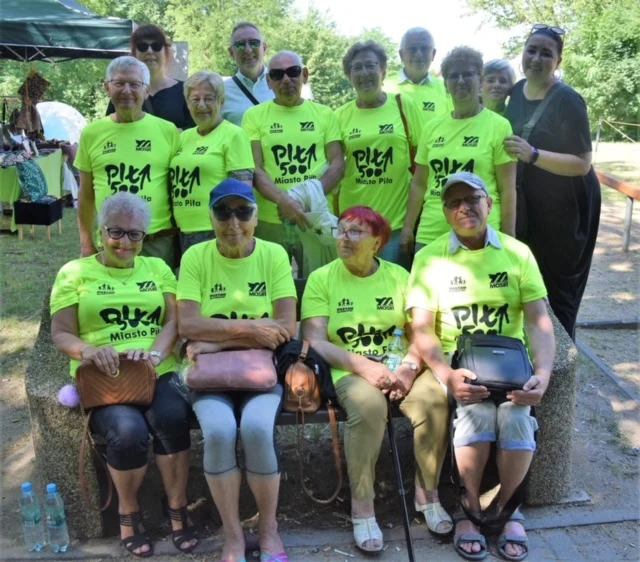 Grupa starszych osób w jaskrawozielonych koszulkach z napisem, siedząca na ławce na tle zieleni, poza nimi kilka osób stoi. Atmosfera jest przyjazna i radosna.