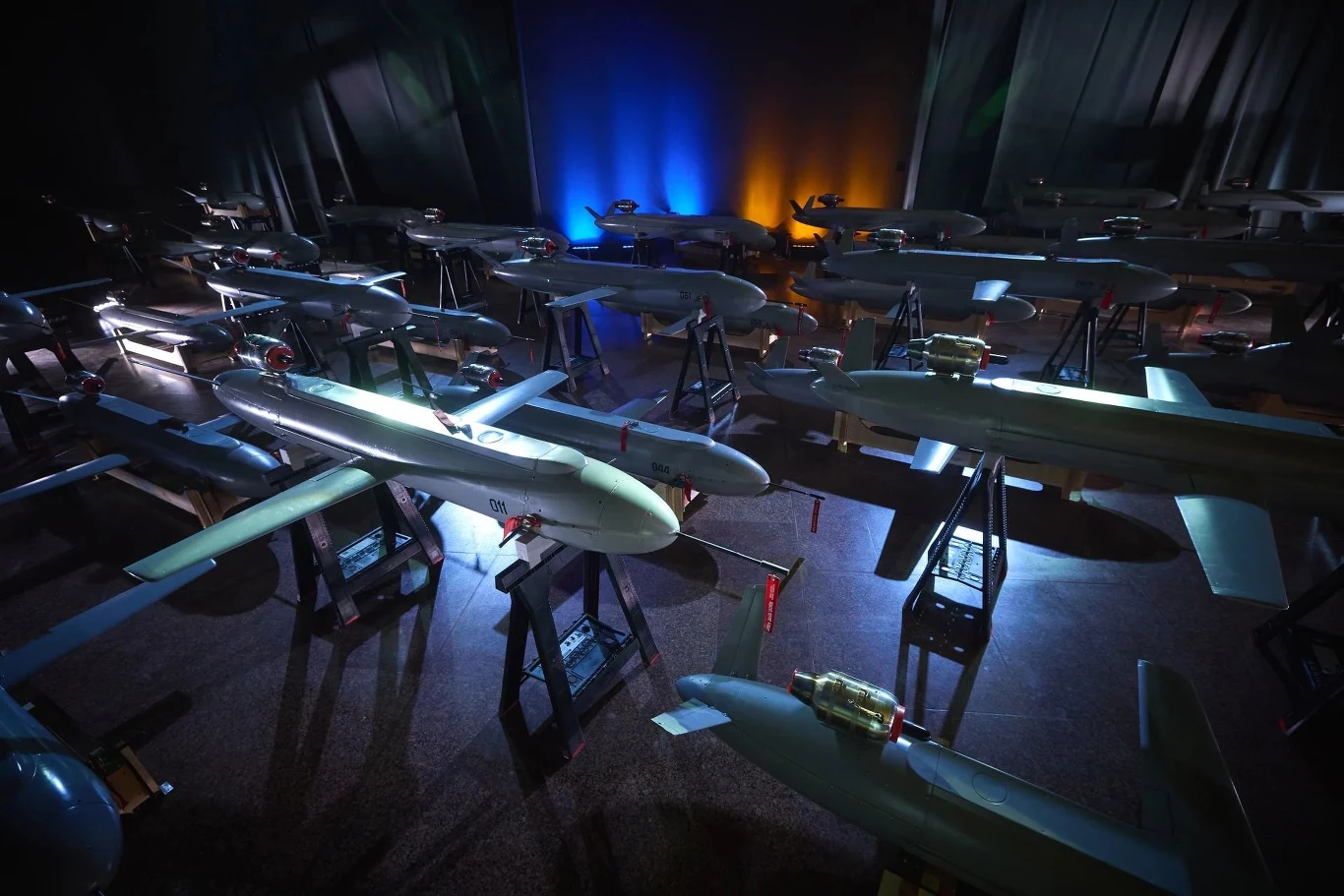 Ukraina prezentuje kolejne drono-rakiety. Było Piekło i Ruta, teraz dołącza Bars Ekspozycja różnorodnych dronów na stojakach w ciemnym pomieszczeniu, oświetlona kolorowymi światłami. Drony ustawione są w rzędach, a tło tworzą zasłony oraz niebiesko-pomarańczowe oświetlenie.