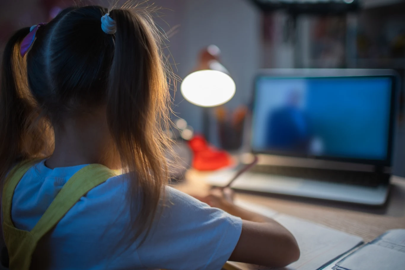 Dziecko poznało w sieci nową osobę. Mężczyzna wykorzystał łatwowierność dziewczynki. Zdj. ilustracyjne. Dziecko z dwoma kucykami siedzi przy biurku w pokoju oświetlonym lampką biurkową. Na biurku znajduje się otwarty laptop, na którym widać rozmyty obraz. Dziecko coś zapisuje na kartce, koncentrując się na ekranie.