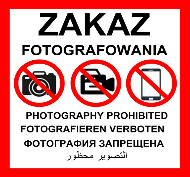 Miejsca oznaczone tym znakiem podlegają zakazowi fotografowania. Znak zakazu fotografowania w kilku językach, przedstawia przekreślone ikony aparatu, kamery wideo i smartfona, wskazując na zakaz używania tych urządzeń. Tekst ostrzegawczy na czerwonym tle.
