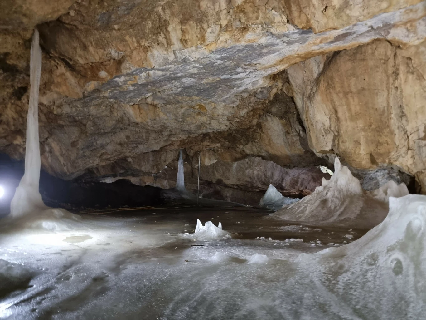 Wnętrze jaskini z widocznymi lodowymi stalagmitami i stalaktytami. Na podłodze oraz ścianach jaskini znajduje się cienka warstwa lodu, co nadaje scenie zimowy wygląd. Skały są naturalnie rzeźbione, tworząc surowy i zimny krajobraz.