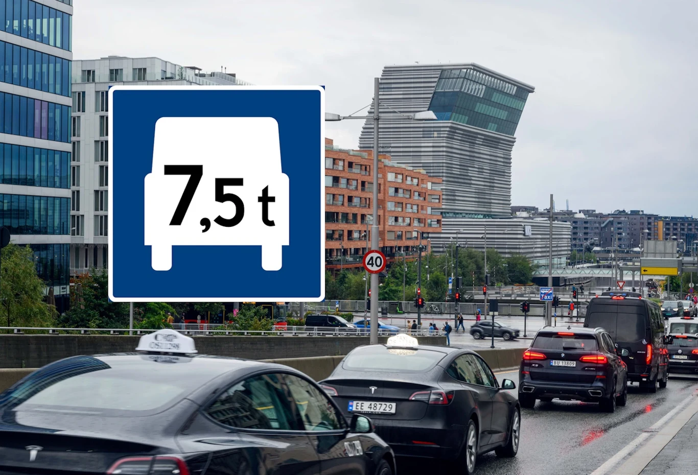 Niebieski znak z literą T zaskakuje kierowców. W Norwegii oznacza pas przeznaczony dla ciężarówek Ruch uliczny w mieście z różnymi samochodami, w tym taksówkami. Widoczny jest znak drogowy o ograniczeniu masy do 7,5 tony. W tle nowoczesne budynki oraz wieżowiec o unikalnej architekturze.