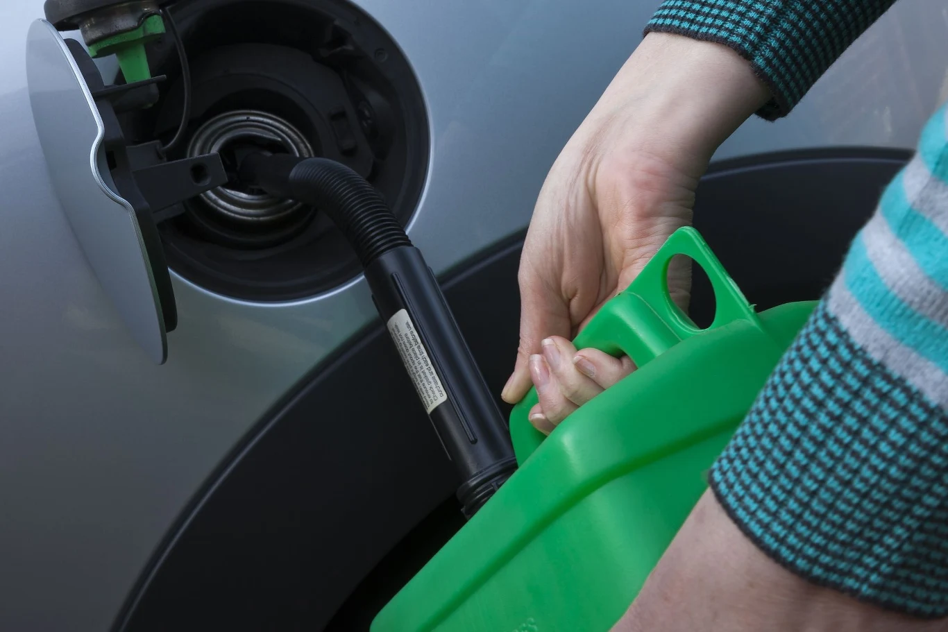 Przepisy określają również litraż naczyń, w których przewożone jest paliwo. Ręce w zielonym swetrze trzymają zielony kanister z paliwem, nalewając je do wlewu paliwa w samochodzie. Wlew samochodu ma otwartą klapę, a ręka trzymająca kanister jest w trakcie przechylania, by wlać paliwo.