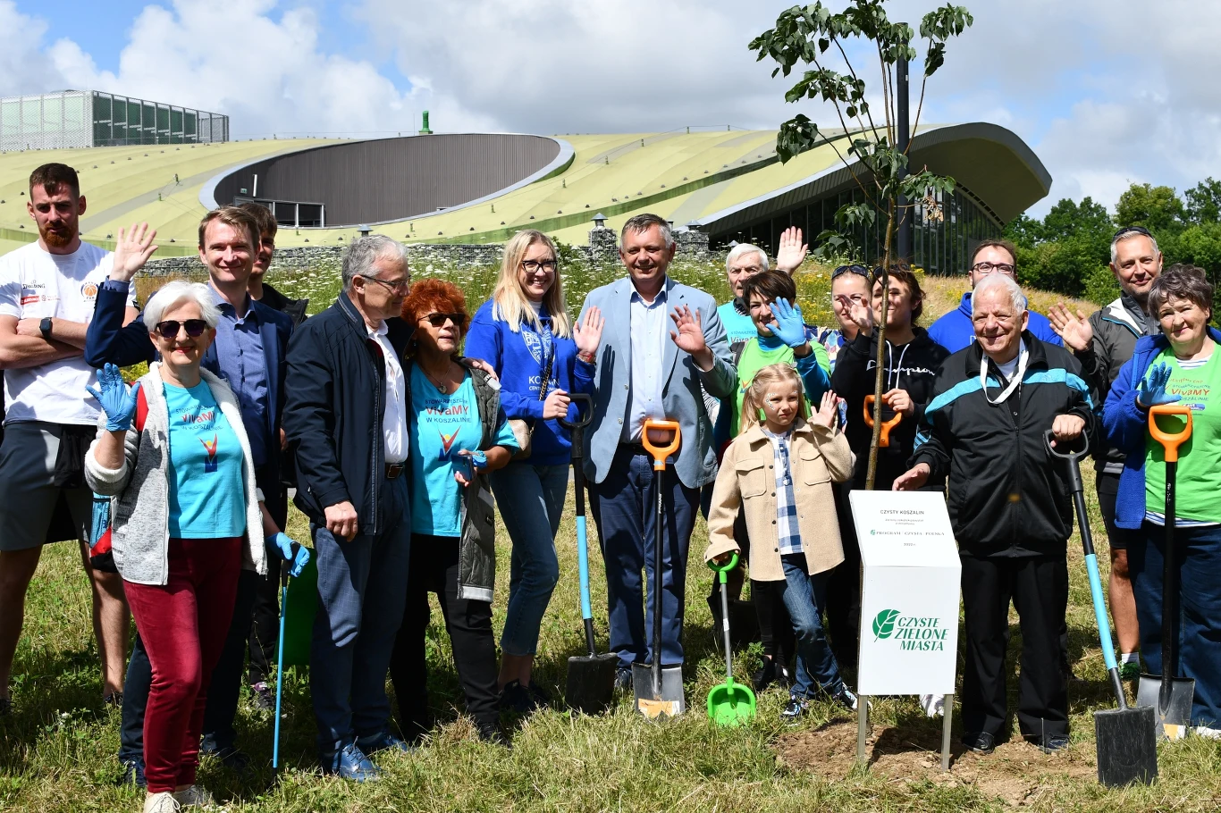 Grupa osób uczestniczących w sadzeniu drzew na tle nowoczesnego budynku z roślinnym dachem. Wszyscy uśmiechnięci, trzymają narzędzia ogrodnicze. Na pierwszym planie młody osobnik dębu z białą tabliczką.