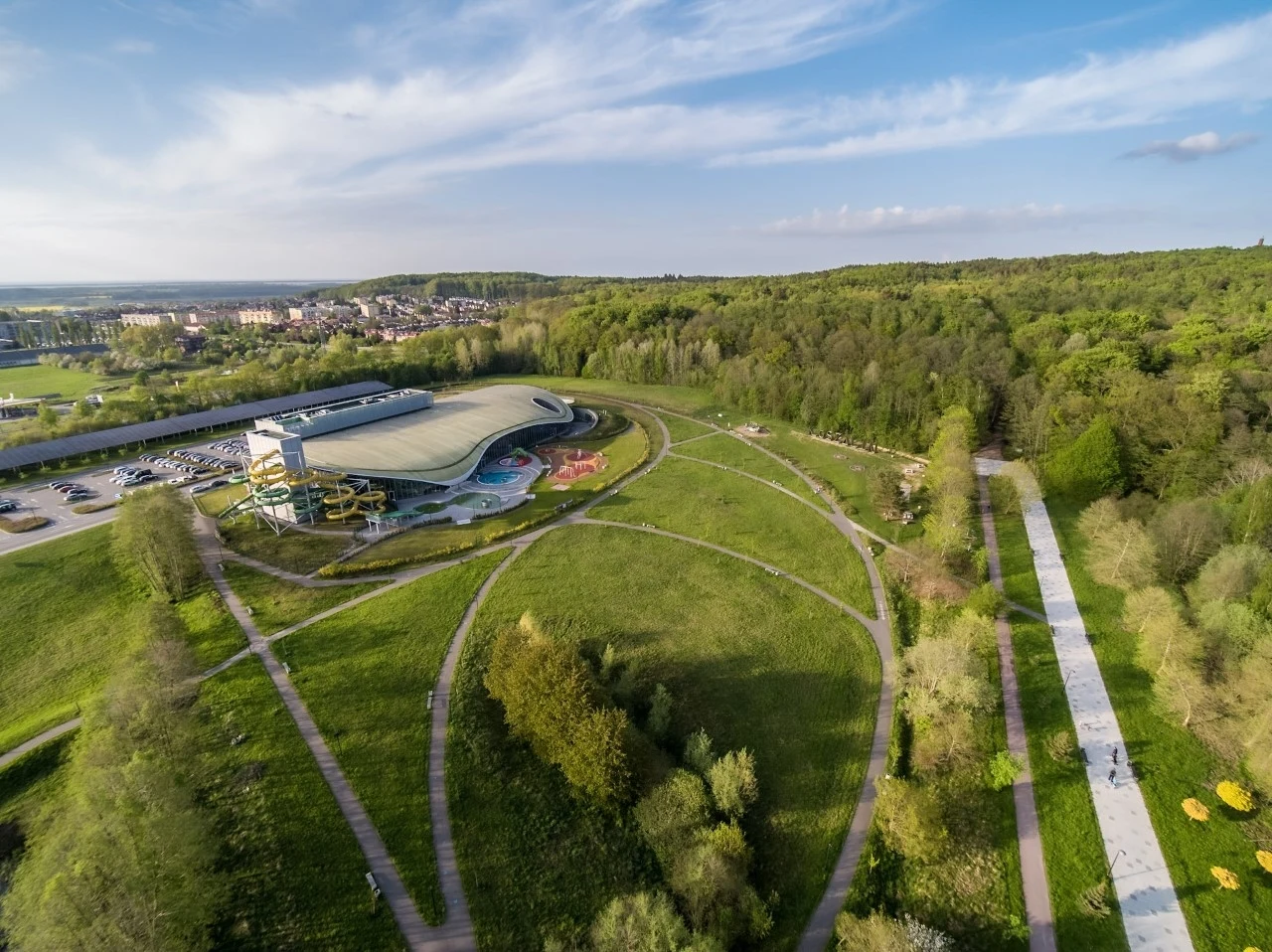 widok z lotu ptaka na kompleks basenowy z zielonymi terenami wokół, ścieżki spacerowe przecinają trawniki, w tle las i rozciągające się miasto