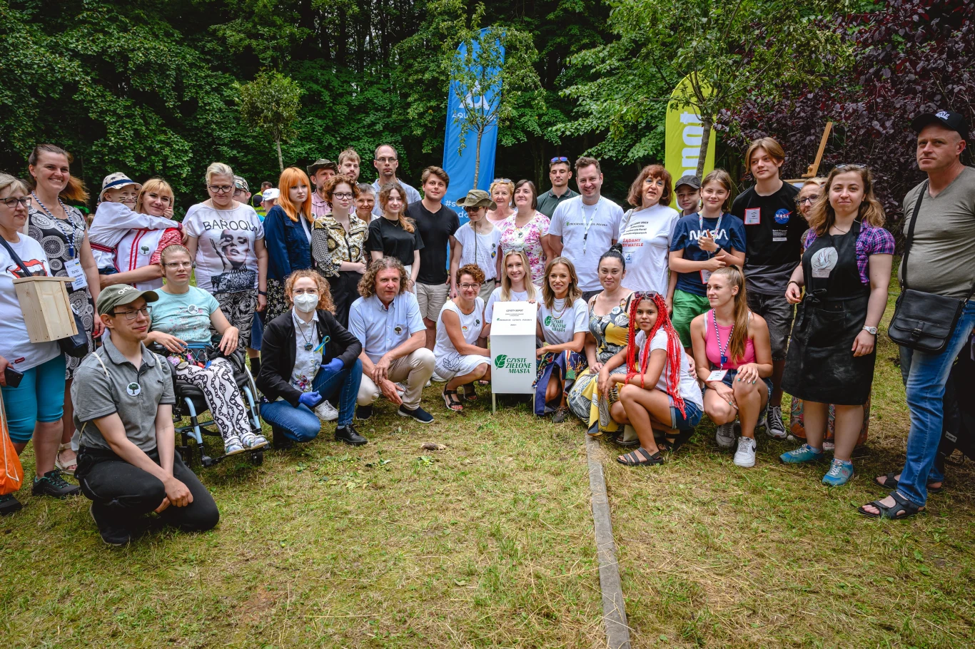 Grupa ludzi różnych płci i wieku pozuje do wspólnego zdjęcia na tle zieleni i banerów. W centrum znajduje się tabliczka informacyjna z logo. Wszyscy są uśmiechnięci i zrelaksowani, sugerując przyjazną atmosferę i wydarzenie integracyjne.
