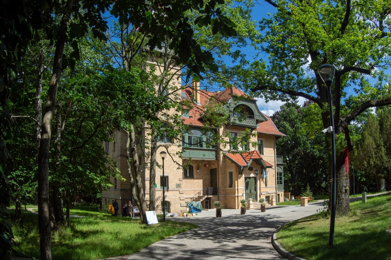 Duży, stylowy budynek z czerwonym dachem otoczony zielonymi drzewami, w jasnym świetle słonecznym, z szeroką ścieżką prowadzącą do głównego wejścia.