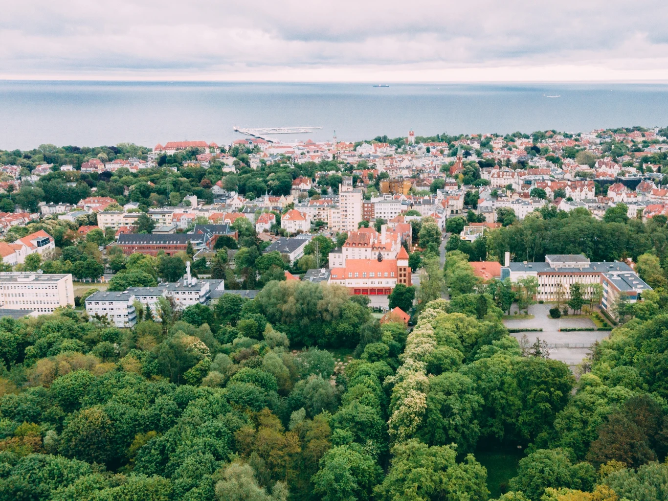 Widok z lotu ptaka na nadmorskie miasto z rozległymi terenami zielonymi i zabudową miejską. W tle widać morze i port, a wokół las i parki. Na horyzoncie znajdują się statki oraz zabudowa mieszkaniowa.