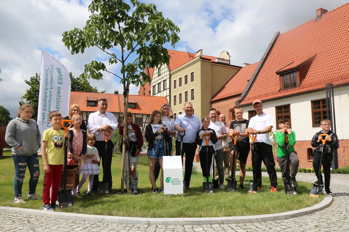 Grupa ludzi w różnym wieku pozuje do zdjęcia na tle budynków z czerwonymi dachami. Na pierwszym planie widoczny jest świeżo zasadzone drzewo. Uczestnicy trzymają w rękach przyrządy ogrodnicze, co sugeruje udział w akcji sadzenia drzew.