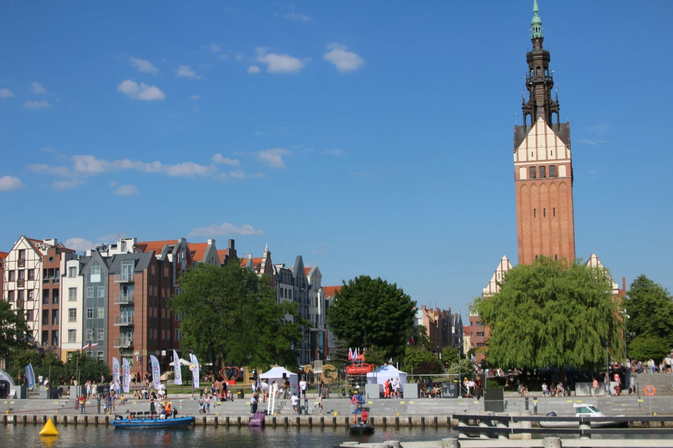 Widok na nadbrzeże z wieżą Bazyliki Mariackiej po prawej stronie, otoczoną zielenią. Po lewej stronie znajdują się kamienice oraz deptak z ludźmi spacerującymi w słoneczny dzień.