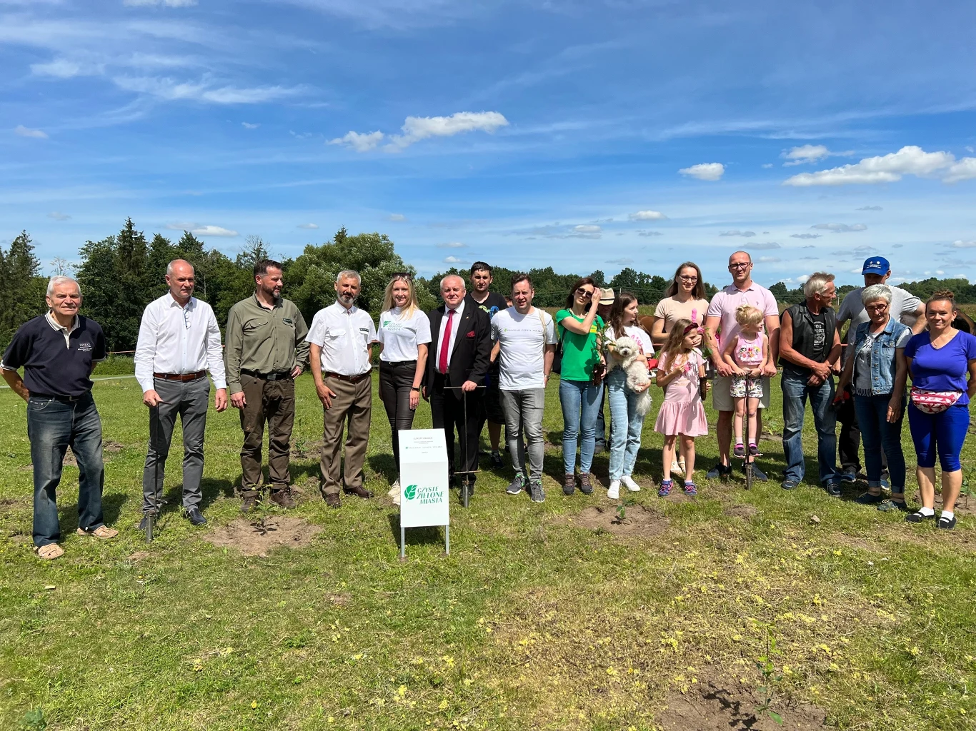 Grupa ludzi pozuje na otwartej przestrzeni, na zielonej trawie pod błękitnym niebem. Stoją obok siebie, niektórzy trzymają dzieci na rękach. Ubrani są w codzienne, nieformalne stroje. W tle widoczny jest las i chmury na niebie.
