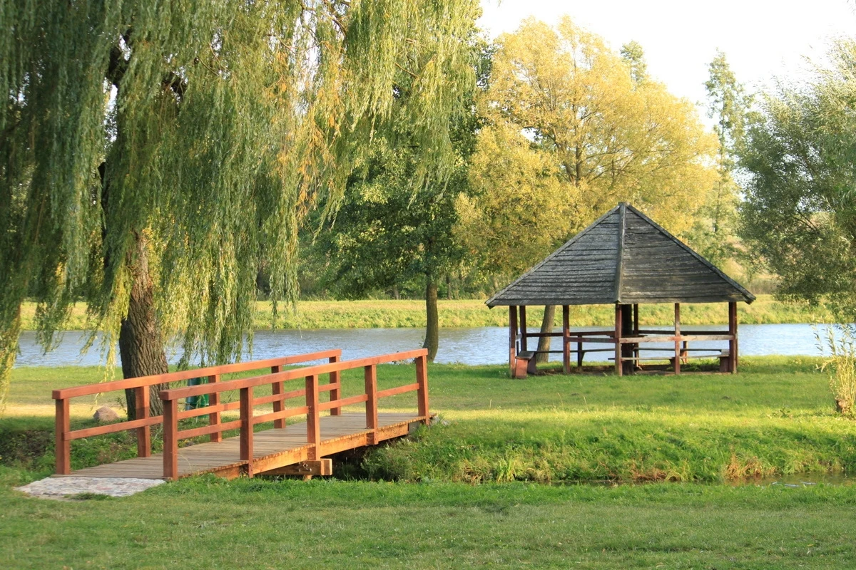Altana ogrodowa przy rzece z drewnianym mostkiem i wierzbą w tle, otoczona zielenią i drzewami.