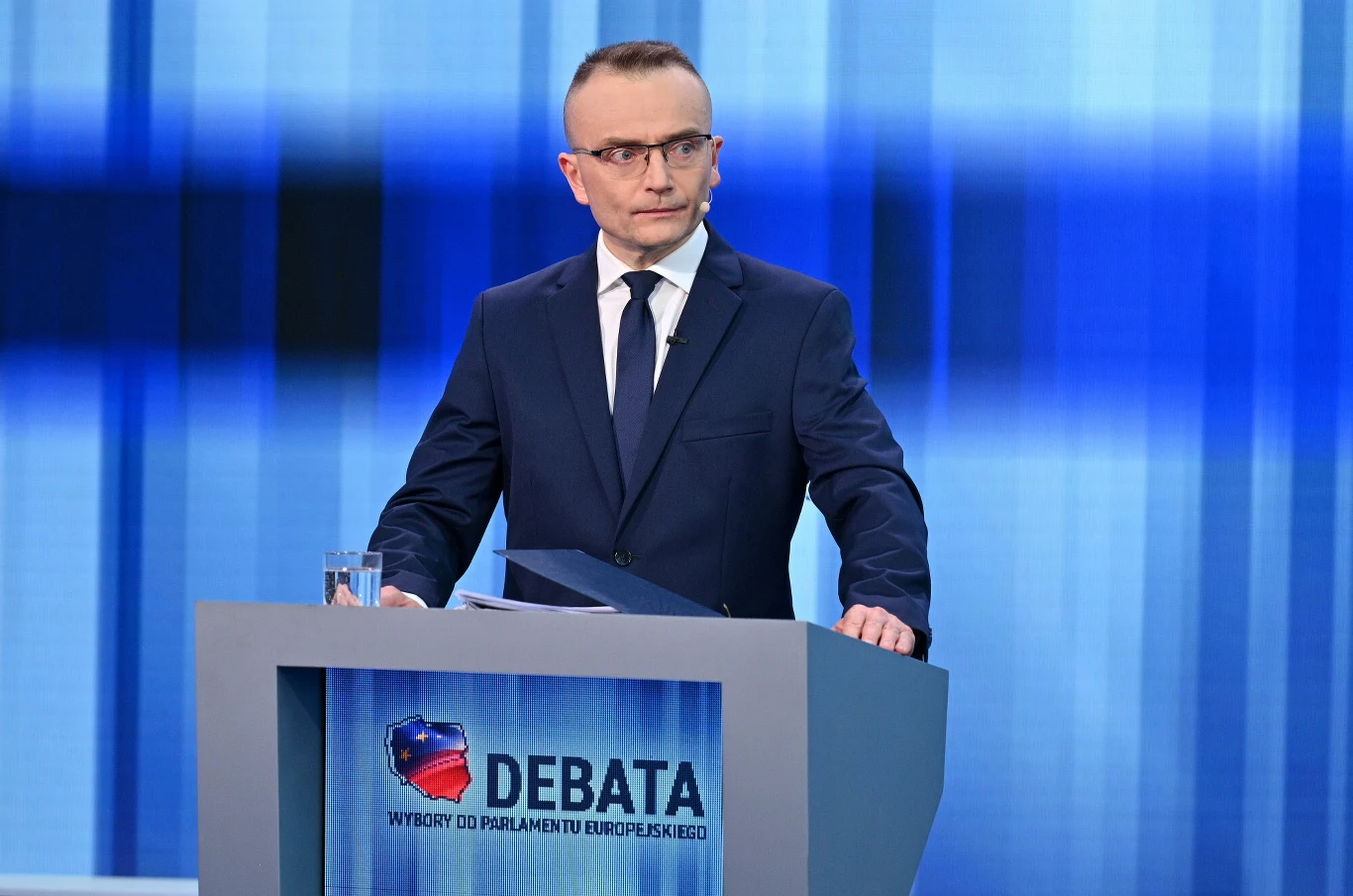 Kandydat na prezydenta Marek Woch Mężczyzna w garniturze, stojący przy mównicy oznaczonej jako "Debata, wybory do Parlamentu Europejskiego". Tło w odcieniach niebieskiego, charakterystyczne dla studia telewizyjnego.