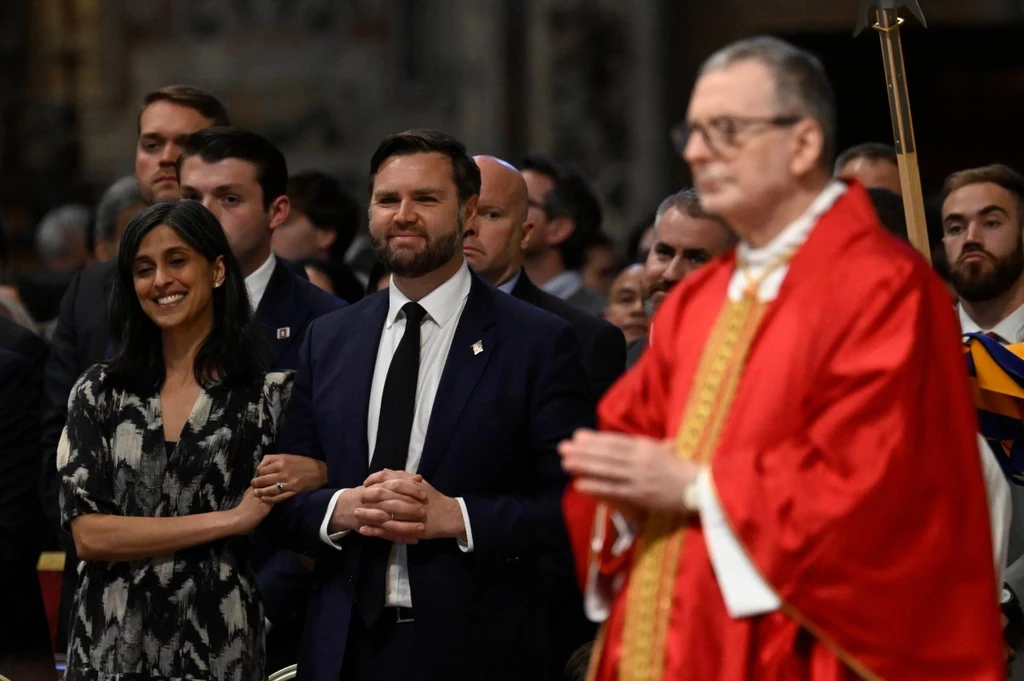 J.D. Vance z żoną Ushą Grupa ludzi w eleganckich strojach w kościele, na pierwszym planie mężczyzna w czerwonych szatach liturgicznych odprawia ceremonię.