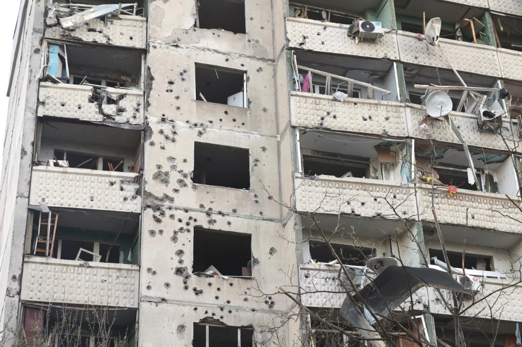 Zniszczony budynek mieszkalny z widocznymi śladami po ostrzale, uszkodzone balkony i okna, potłuczone szyby oraz porozrzucane elementy wyposażenia wewnętrznego.