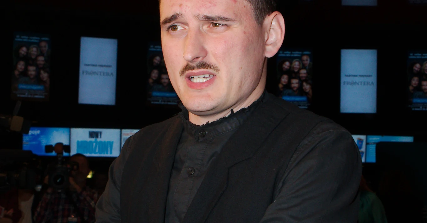Dawid Podsiadło ze szczerym wyznaniem Mężczyzna w ciemnym stroju z wąsami stoi na tle ekranów z plakatami filmowymi.