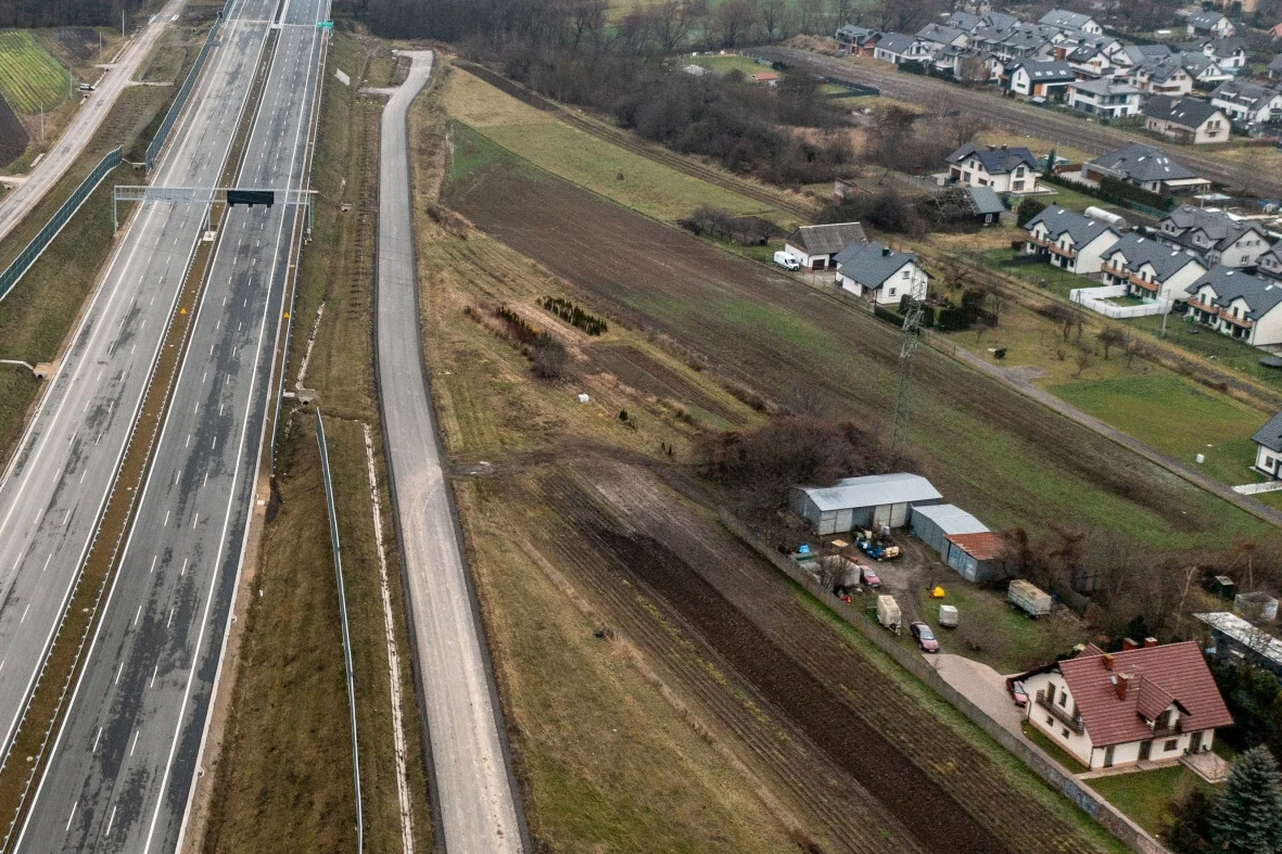 Część osób mieszkających w pobliżu Północnej Obwodnicy Krakowa skarży się na ciągły hałas związany z brakiem ekranów dźwiękochłonnych Autostrada biegnąca wzdłuż polnych terenów, z domami mieszkalnymi po jednej stronie. Widoczne pola uprawne i grupy domów w tle. Tereny są uporządkowane, a infrastruktura drogowa jest nowoczesna.