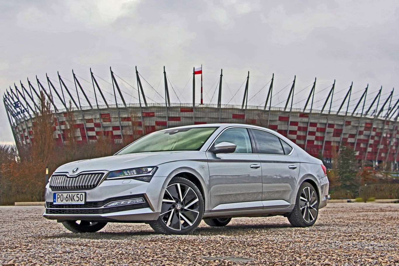 Skoda Superb III - czy warto ją kupić, jaki silnik wybrać? Srebrny samochód osobowy zaparkowany przed Stadionem Narodowym w Warszawie, dominującym w tle.