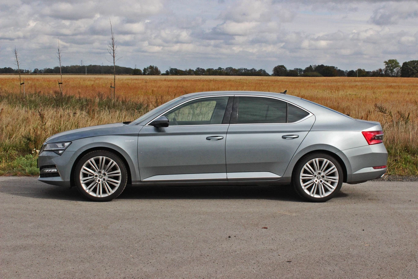 Skoda Superb III ma 486 cm długości Srebrny samochód osobowy zaparkowany przy drodze, z polami w tle i lekkim zachmurzeniem na niebie.