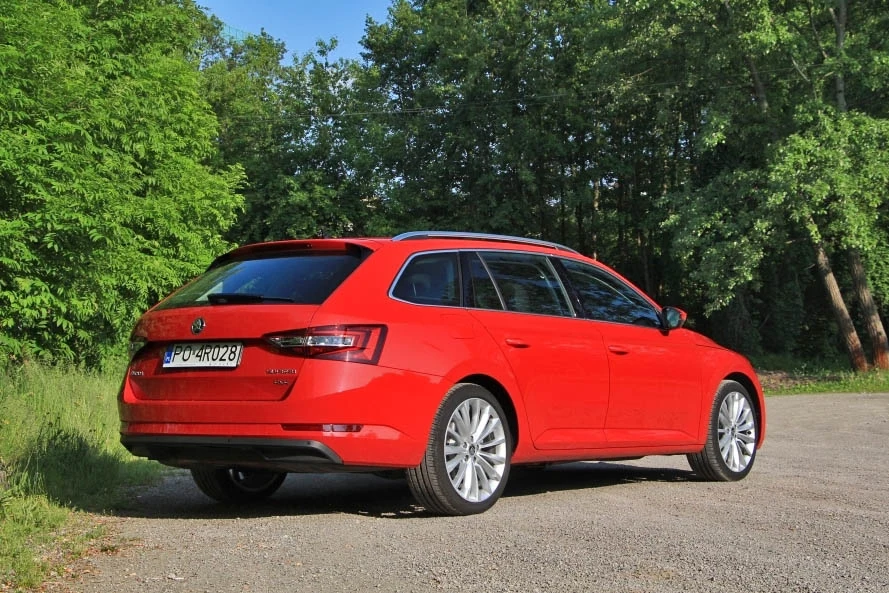 Skoda Superb III dostępna była jako liftback oraz kombi Czerwony samochód kombi marki Škoda zaparkowany tyłem na żwirowej drodze, otoczony bujną zielenią lasu.