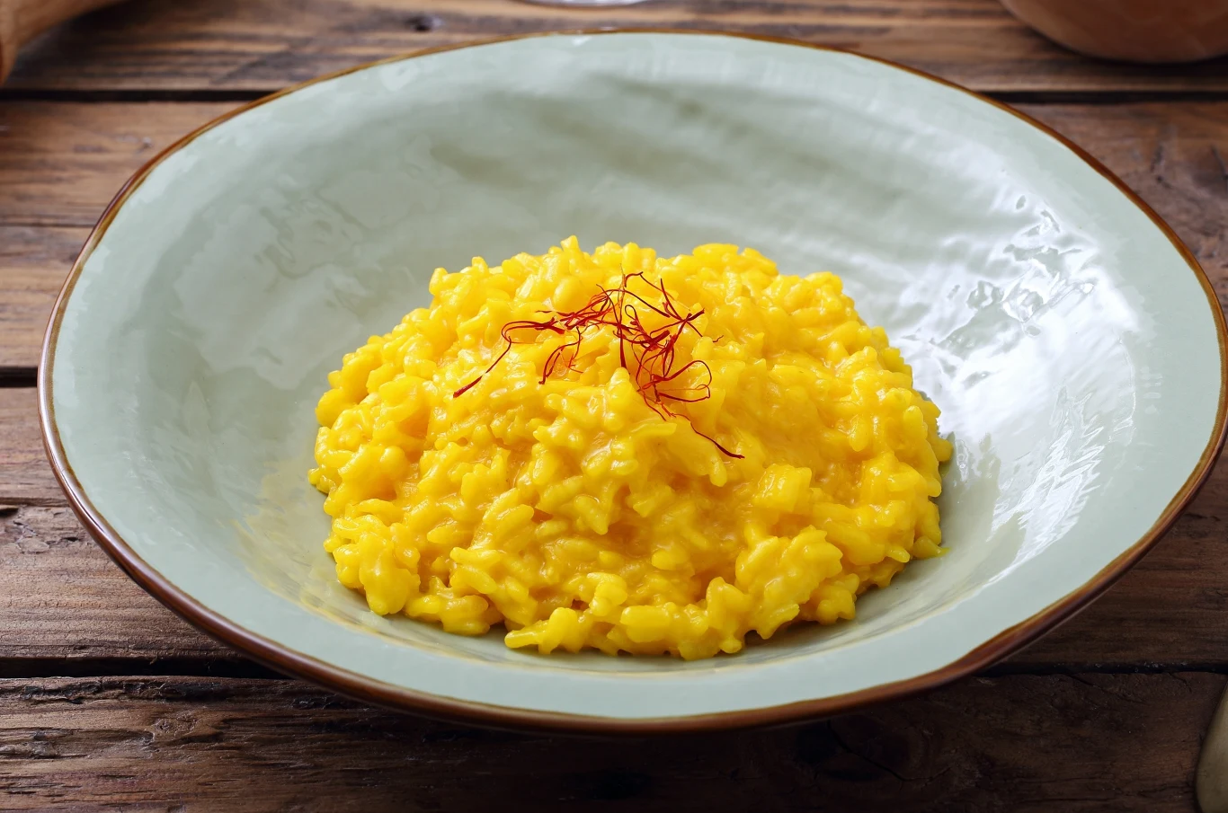 Jasnożółte risotto na szerokim, szarym talerzu. Dekoracja z kilkoma nitkami szafranu na wierzchu. 