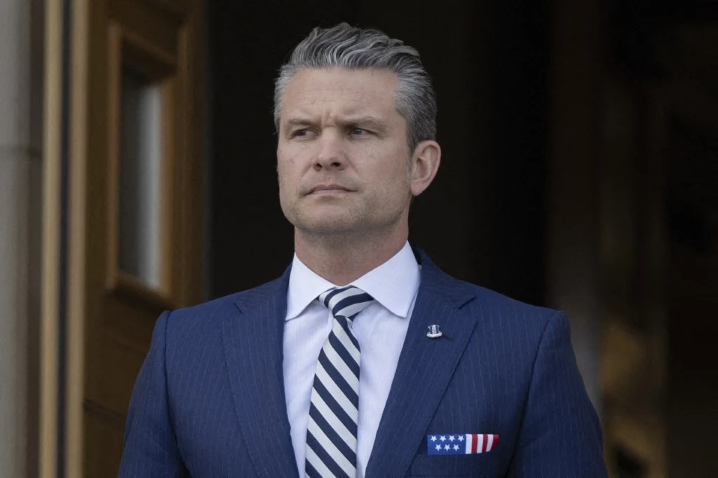 USA. Pete Hegseth traci kolejnego współpracownika Mężczyzna w eleganckim garniturze i krawacie w paski, stojący przed wejściem do budynku. Ma krótko przystrzyżone siwe włosy i patrzy przed siebie. Na klapie widoczna jest przypinka, a w brustaszy chusteczka z motywem amerykańskiej flagi.