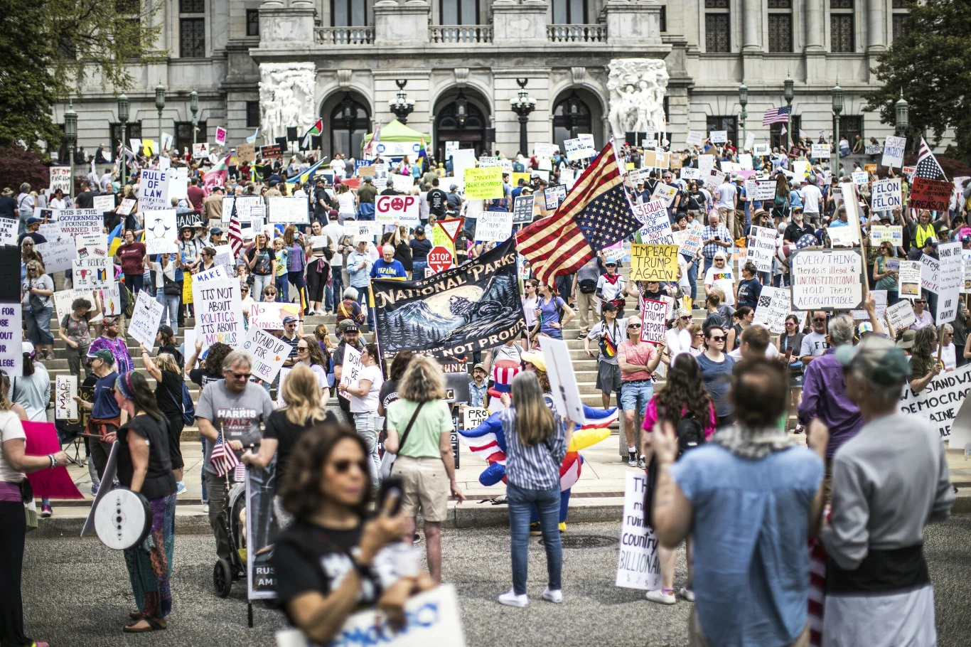 Amerykanie protestowali w całym kraju Tłum ludzi z transparentami i flagami przed dużym budynkiem rządowym. Uczestnicy trzymają różnorodne hasła dotyczące tematyki politycznej i społecznej. Na pierwszym planie widoczne są osoby robiące zdjęcia i rejestrujące wydarzenie.