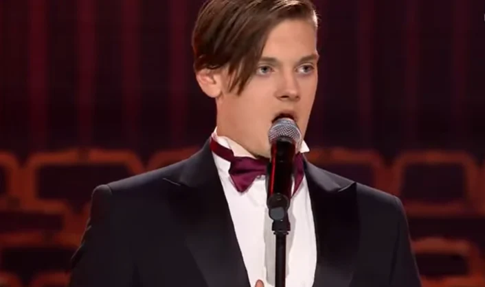 Krystian Leśnik zdobył Platynowy Przycisk Młody mężczyzna śpiewający do mikrofonu, ubrany w elegancki garnitur i muszkę, na tle rozmytego czerwonego tła.