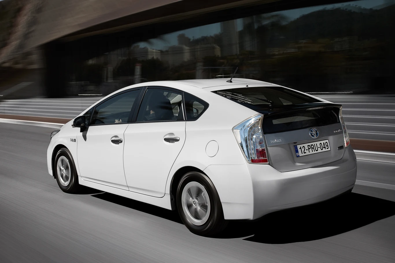 Jadący po ulicy biały samochód osobowy marki Toyota Prius, widok z tyłu. Tło rozmazane przez ruch, co sugeruje prędkość pojazdu.