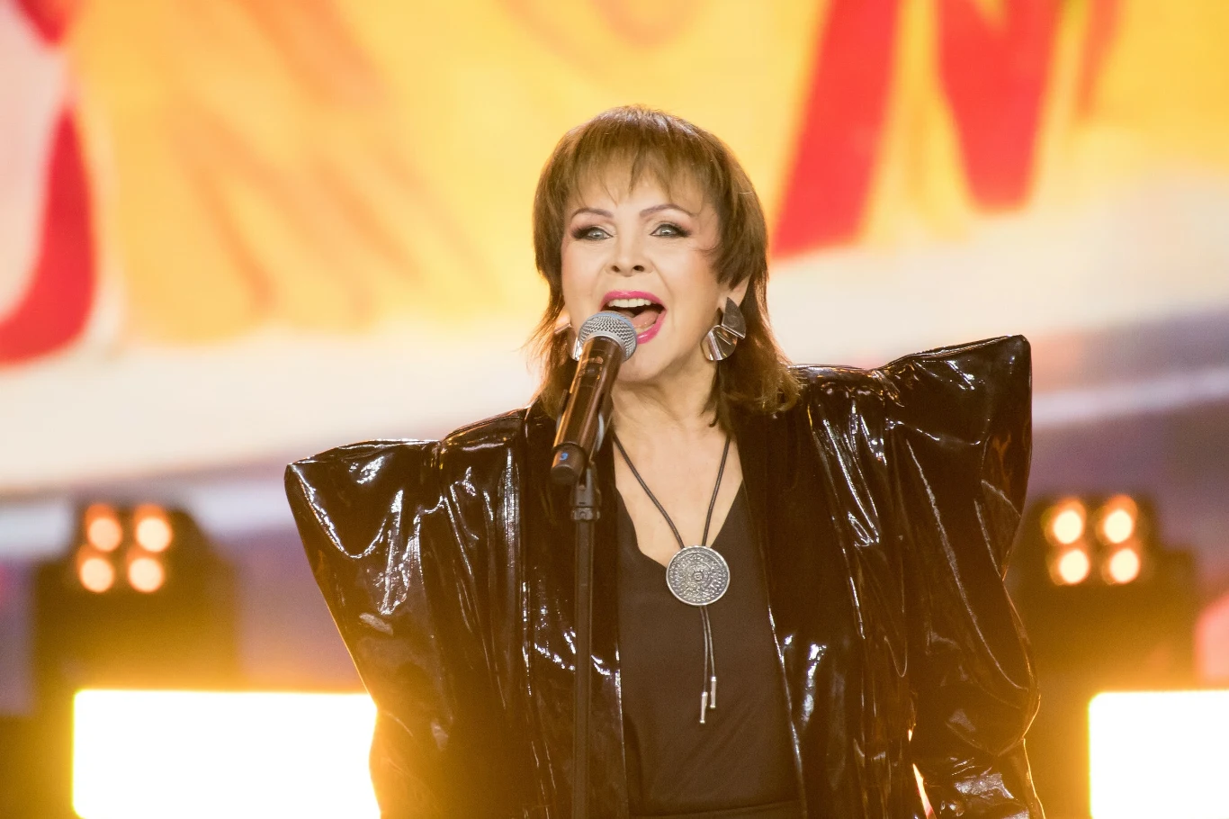 Izabela Trojanowska jest legendą polskiej sceny Osoba śpiewająca na scenie z mikrofonem w ręku, ubrana w czarny strój z charakterystycznymi szerokimi ramionami, za nią jasne oświetlenie sceniczne i kolorowe tło.