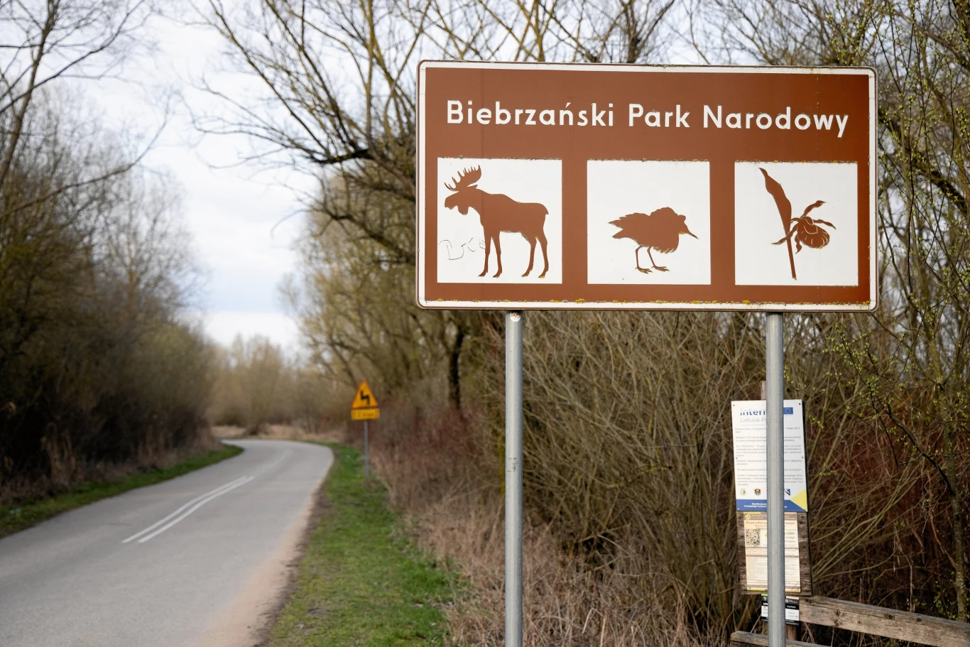 Droga prowadząca do Biebrzańskiego Parku Narodowego z umieszczonymi znakami informacyjnymi przedstawiającymi łosia, ptaka i ważkę. W tle widać zarośla i drzewa typowe dla obszaru parku.
